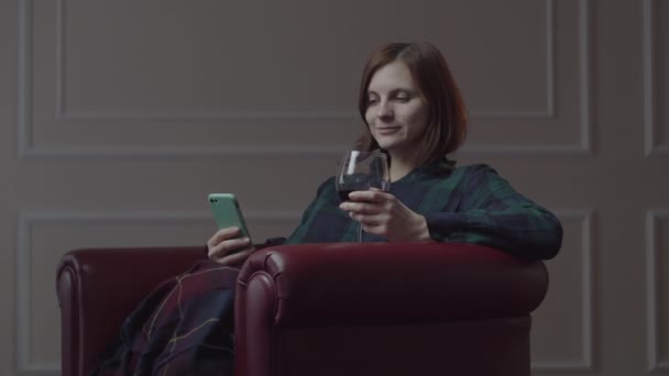 Молода 30 років жінка п'є червоне вино і використовує мобільний телефон, сидячи в затишному червоному кріслі вдома. Жінка сама з мобільним телефоном і келихом вина . — стокове відео