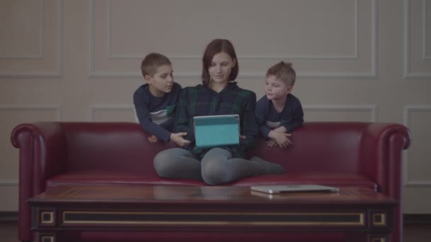 Joven madre de 30 años sentada en el sofá y usando tableta, dos chicos con la misma ropa viendo gadget con mamá. Familia viendo contenido en línea en casa . — Vídeo de stock