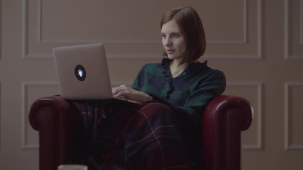 30 'lu yaşlarda, dizüstü bilgisayarda yazan genç bir kadın rahat koltukta ekose kumaşla oturuyor. Evde tek başına çalışan bir kadın.. — Stok video