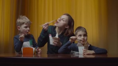 İki çocuklu bir anne evde yemek çubuklarıyla Asya yemeği yiyor. Mutlu bir aile, teslimat kutusundan yemek yiyor.. 