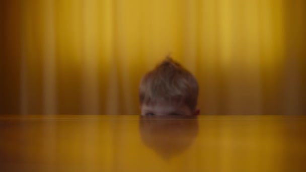 Portret van een blonde kleuter die glimlacht en naar de camera kijkt. Kinderen gaan thuis op de tafel liggen. Glimlachend kind toont duimen omhoog. — Stockvideo