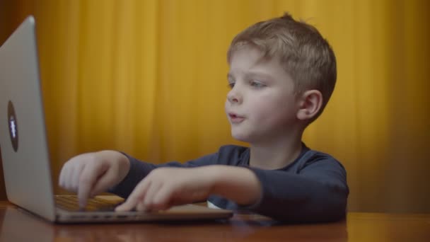 Biondo bambino in età prescolare digitando sul computer portatile e sorridente guardando lo schermo del computer. Bambino che impara online a casa seduto a tavola . — Video Stock