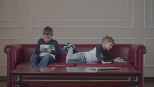 Dva sourozenci ve stejném oblečení, kteří používají gadgets a hrají si na gauči. Jeden kluk bere svému bratrovi mobil.. — Stock video