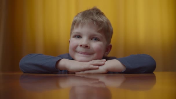 Porträtt av blonda förskolan pojke ler och tittar till kameran. Ungar går upp på bordet hemma. Leende unge visar tummen upp. — Stockvideo