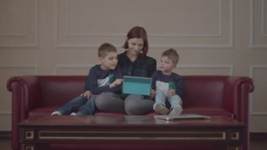 30 'lu yaşlarda genç bir anne kanepede oturuyor ve tablet bilgisayar kullanıyor. Aynı kıyafetleri giymiş iki çocuk annemle birlikte cihaz izliyor. Aile evde çevrimiçi içeriği izliyor. 
