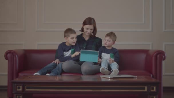 若い30代の母親がソファに座ってタブレットコンピュータを使用して、同じ服を着た2人の男の子が母親とガジェットを見ています。家庭でオンラインコンテンツを見ている家族. — ストック動画