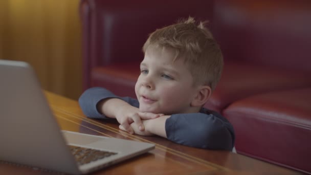 Rubia preescolar niño escribiendo en el ordenador portátil y sonriendo mirando a la pantalla del ordenador. Aprendizaje de niños en línea en casa sentado en la mesa . — Vídeos de Stock