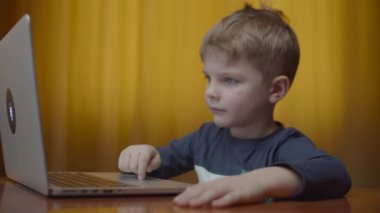 Bilgisayar ekranına bakıp gülümseyen sarışın anaokulu öğrencisi. Çocuk evde, masada oturarak öğreniyor.. 