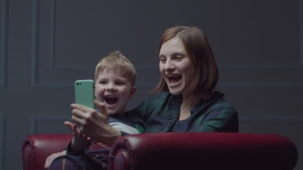 Joven madre de 30 años con hijo preescolar utilizando el teléfono móvil, sentado en un sillón acogedor. Familia divirtiéndose con gadget en casa . — Vídeos de Stock