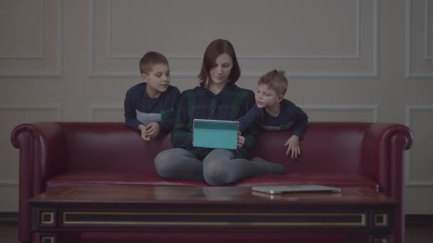 Ung 30-tals mamma sitter på soffan och använder surfplatta dator, två pojkar i samma kläder tittar på gadget med mamma. Familj titta på online-innehåll hemma. — Stockvideo