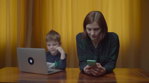 Menino pré-escolar loiro digitando no laptop e na mãe com telefone celular sentado na mesa em casa. Kid aprendendo online, mãe ajudando-o . — Vídeo de Stock