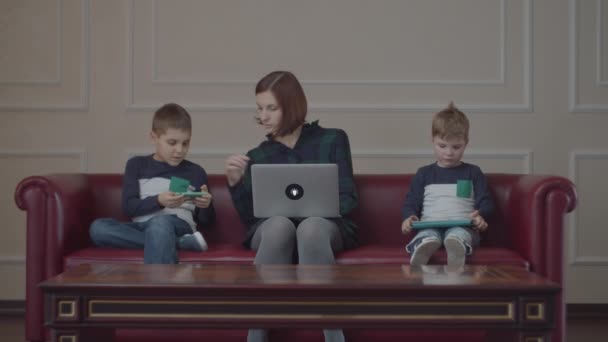 Giovane madre di 30 anni con computer portatile e due ragazzi con gli stessi vestiti usando gadget, seduti sul divano. Famiglia che guarda contenuti online su diversi dispositivi a casa . — Video Stock