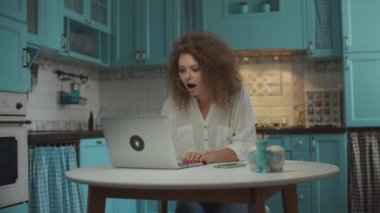 Genç kıvırcık saçlı 20 'li yaşlarda bir kadın laptopta iyi haberler okuyor ve çok heyecanlanıyor, yumruklarını başarıyla kaldırıp ellerini çırpıyor, mavi mutfaktaki masada oturuyor.. 