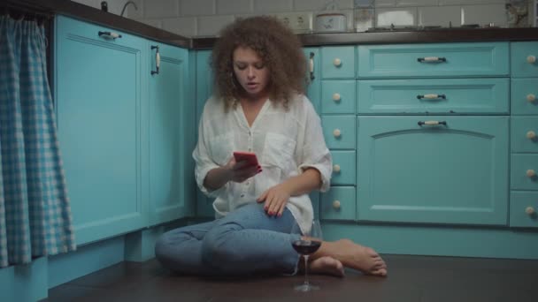 Unga lockigt 20-tal upprörd kvinna läser dåliga nyheter på mobiltelefon och dricka glas rött vin sitter på köksgolvet. Kvinna med vin och pryl hemma. — Stockvideo