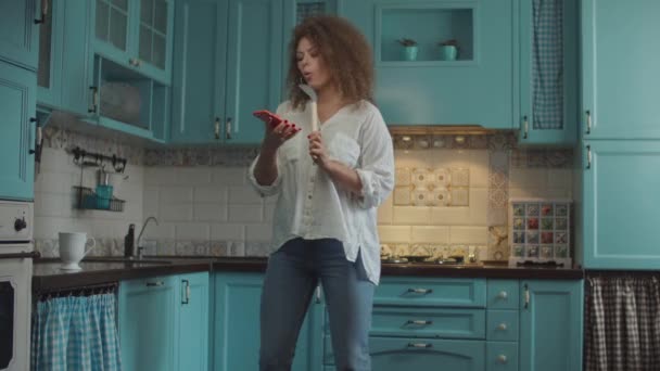 Sorrindo encaracolado 20s mulher em jeans dançando alegremente com colher como microfone e telefone celular em mãos em casa cozinha azul . — Vídeo de Stock