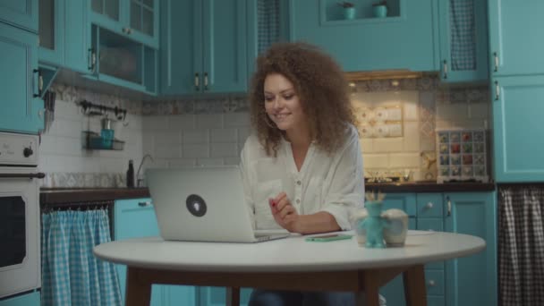年轻的卷发20多岁的女人坐在蓝色厨房的桌子旁，用笔记本电脑完成了工作，并满意地举起双手. — 图库视频影像