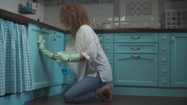 20-an muda perempuan keriting dalam membersihkan sarung tangan penyemprotan deterjen dan menyeka lemari dapur dengan kain. Wanita lelah membuat rumah bersih-bersih . — Stok Video