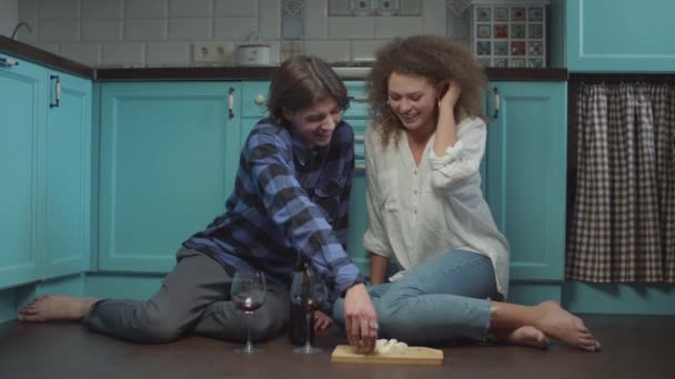 Mladý dvacetiletý pár pije víno a jí sýr na podlaze v kuchyni. Šťastný usměvavý muž a žena slaví s sklenicemi vína doma. — Stock video