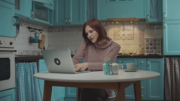 Young 30s woman talking by mobile phone emotionally with hands gesticulating, typing on laptop, sitting at the table on home blue kitchen. — Stock Video