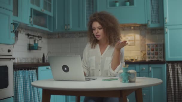젊은 곱슬머리 20 대 여성 이 부엌에서 비디오 통화를 하고 있습니다. 여성 이 컴퓨터 화면에 감정적으로 이야기하고 손으로 몸짓을 하며 집 식탁에 앉아 — 비디오