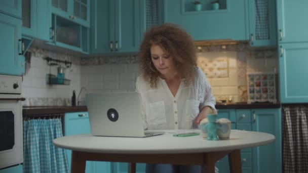 Young curly 20s working female looking through paper documents with charts and verifying information with laptop, sitting on cozy home blue kitchen. — Stock Video