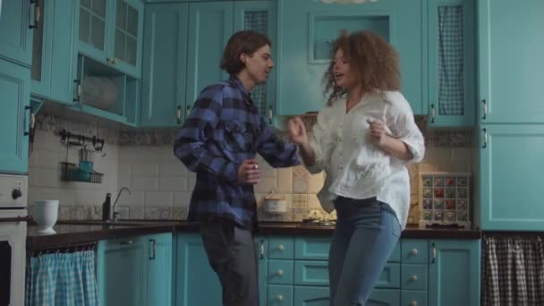 Joven pareja feliz de 20 años bailando en ropa casual con utensilios de cocina en las manos en la acogedora cocina azul. Hombre y mujer divirtiéndose con rodillo y cucharada en la cocina casera — Vídeo de stock