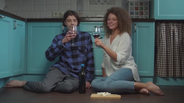 Pareja joven de 20 años bebiendo vino y comiendo queso sentado en el suelo de la cocina. Feliz hombre y mujer sonrientes celebrando con copas de vino en casa . — Vídeo de stock