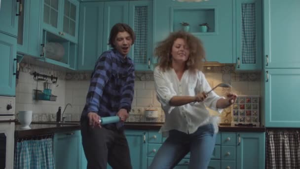 Young happy 20s couple dancing in casual clothing with kitchenware in hands on cozy blue kitchen. Man and woman having fun with rolling pin and scoop at home kitchen — Stock Video