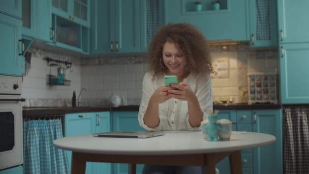 Jovem de 20 anos encaracolado surfando online usando telefone celular e rindo sentado à mesa na cozinha azul. Mulher com celular nas mãos em casa . — Vídeo de Stock