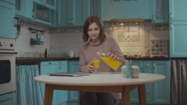 Mujer joven de 30 años que vierte y bebe jugo de naranja, mirando a la computadora portátil sentada en la mesa en la cocina azul. Mujer trabajadora en casa . — Vídeos de Stock