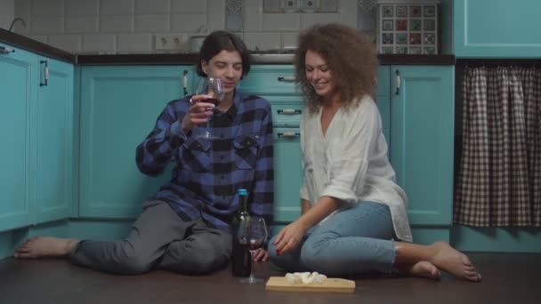 Pareja joven de 20 años bebiendo vino y comiendo queso sentado en el suelo de la cocina. Feliz hombre y mujer sonrientes celebrando con copas de vino en casa . — Vídeo de stock