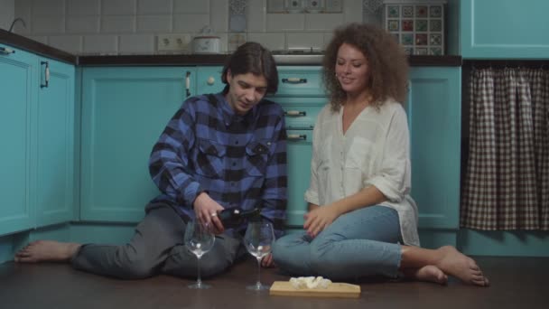 Pareja joven de 20 años bebiendo vino y comiendo queso sentado en el suelo de la cocina. Feliz hombre y mujer sonrientes celebrando con copas de vino en casa . — Vídeos de Stock