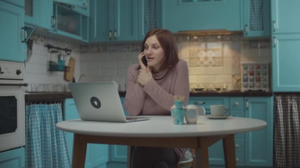Молодая 30-летняя женщина в хорошем настроении говорит по мобильному телефону эмоционально с жестикулирующими руками, сидя за столом с ноутбуком на домашней голубой кухне . — стоковое видео