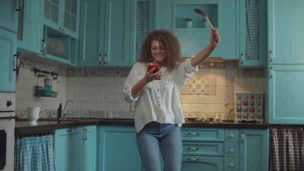 Souriant frisé 20s femme en jeans dansant joyeusement avec scoop comme microphone et téléphone portable dans les mains sur la maison cuisine bleue . — Video