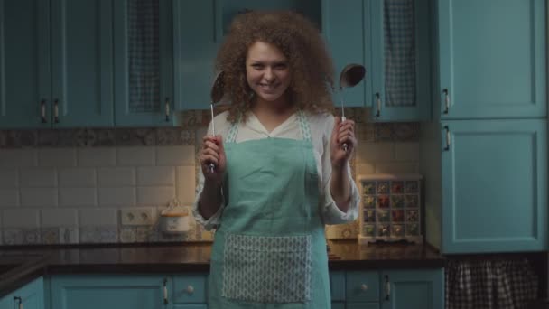 Joven cabello rizado 20s mujer en delantal con utensilio de cocina en las manos de pie en la cocina azul y mover las manos de manera divertida. Mujer con cucharadas en las manos sonriendo mirando a la cámara . — Vídeos de Stock