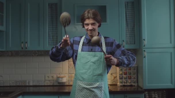 Jovem morena homem de 20 anos em avental com utensílio de cozinha em mãos em pé na cozinha azul e mãos móveis de forma engraçada. Homem com colher nas mãos sorrindo olhando para a câmera . — Vídeo de Stock