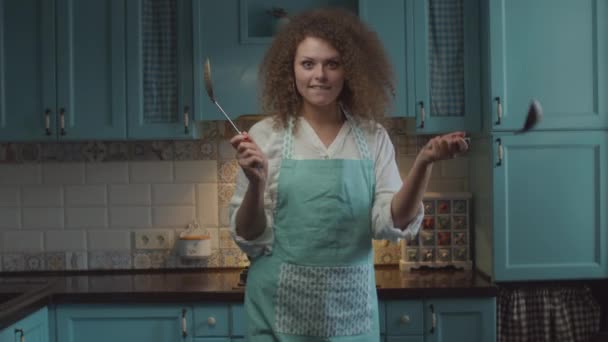 Fiatal göndör haj 20-as nő kötényben konyhai eszközökkel a kezében áll a kék konyha és mozgó kezét vicces módon. Nő gombóccal a kezében mosolyog, kamerába néz.. — Stock videók