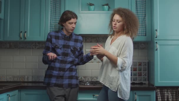 Молода пара 20-х років свариться, стоячи на синій кухні вдома, дівчина показує мобільний телефон і кричить звинувачуючи хлопця. Сімейний ревнивий конфлікт . — стокове відео