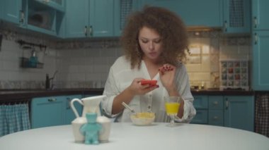 20 'li yaşlarda kıvırcık saçlı bir kadın kahvaltısını tek başına yapıyor ve elinde cep telefonu ile mutfakta bekliyor. Kadın yemek fotoğrafı çekiyor ve telefon ekranı arıyor. Evde yemek masasında oturuyor.. 