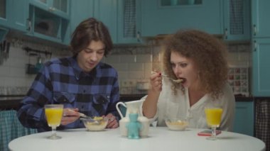 Genç mutlu 20 'li yaşlardaki çift rahat mavi mutfakta kahvaltı yapıyor. Erkek ve kadın mısır gevreğini vejetaryen sütüyle yiyor ve evde ağır çekimde portakal suyu içiyor.. 