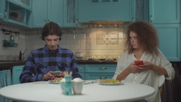 Jeune couple 20s phubbing l'un l'autre tout en dînant avec des gadgets sur cuisine bleue. Homme et femme mangeant des repas et utilisant des téléphones portables à la maison au ralenti . — Video
