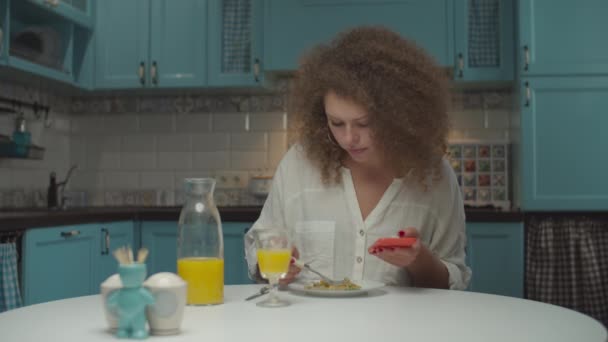 Joven mujer de 20 años pelo rizado comiendo comida sola en la cocina. Mujer con teléfono móvil en las manos cenando en casa . — Vídeos de Stock