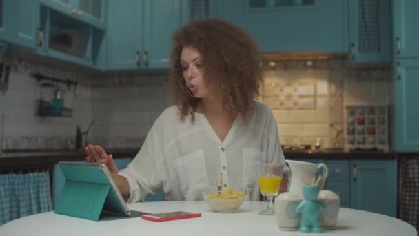 Joven mujer de 20 años cabello rizado desayunando solo y de compras en línea en la tableta de la cocina azul. Mujer haciendo la compra con tarjeta de crédito en línea mientras come en casa . — Vídeo de stock