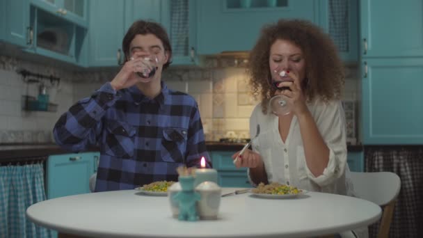 Jonge gelukkige twintigers echtpaar in casual kleding drinken rode wijn met diner op gezellige blauwe keuken. Man en vrouw die thuis in slow motion eten en wijn drinken. — Stockvideo