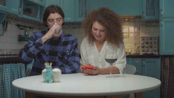 Jonge twintiger jaren koppel drinken rode wijn met gadget op blauwe keuken. Man en vrouw kijken naar mobiele telefoon in vrouwen handen met interesse tijdens het drinken van wijn thuis in slow motion. — Stockvideo