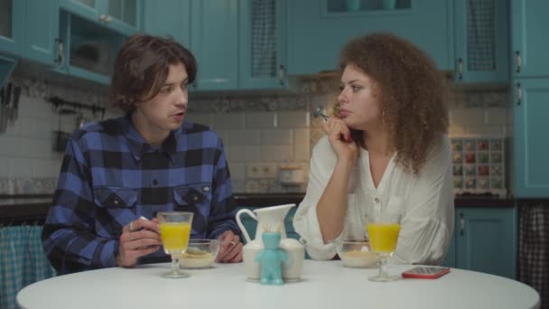 Unga glada 20-talet par i casual kläder som äter frukost på mysigt blått kök. Man och kvinna äter majsflingor med vegansk mjölk och dricker apelsinjuice hemma i slow motion. — Stockvideo
