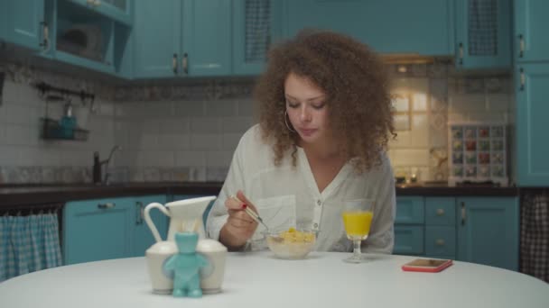 Joven mujer de 20 años cabello rizado desayunando sola, sosteniendo el teléfono móvil en las manos en la cocina. Hembra comiendo copos de maíz con leche vegana sentada en la mesa de comedor en casa . — Vídeos de Stock