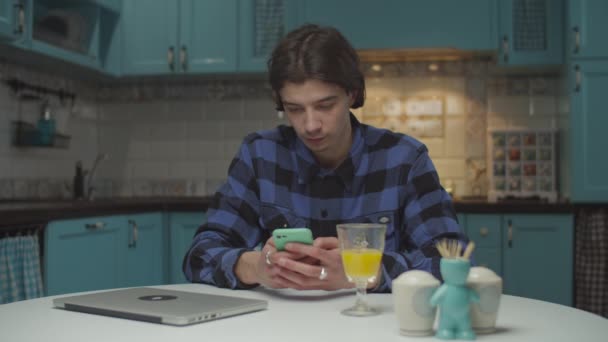 Hombre joven de 20 años navegando en línea utilizando el teléfono móvil y sonriendo sentado en la mesa de comedor en la cocina azul. Hombre con teléfono celular en las manos en casa . — Vídeo de stock