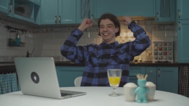 Junger Mann in den 20er Jahren liest gute Nachrichten auf Laptop und wird sehr aufregend, reckt triumphierend die Fäuste, sitzt am Tisch auf blauer Küche. — Stockvideo