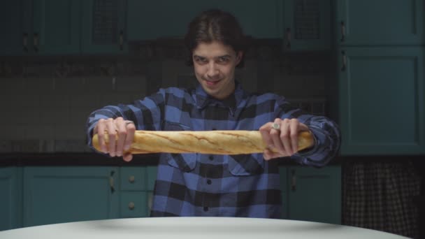 Hombre joven de 20 años comiendo baguette de manera divertida en la cocina. Hombre partiendo el pan mirando a la cámara y sonriendo. Divertido comer . — Vídeos de Stock